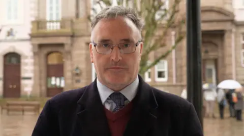 Patrick Lynch is standing in the street and is looking serious. He has short grey hair and is wearing glasses. He is wearing a smart plum-coloured woollen coat and a white and blue checked shirt with navy and white spotted tie and a deep red jumper. There are people holding umbrellas behind him.