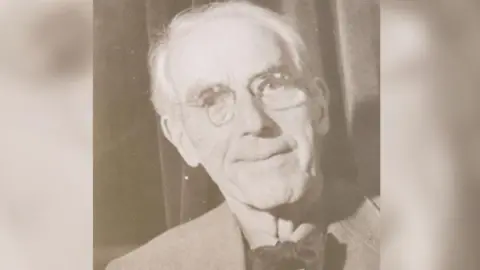 South West Heritage Trust Photographer Henry Wykes who documented Exeter's history is photographed aged 87. He is wearing a suit jacket with a bow tie and glasses in this head and shoulders shot and is smiling at the camera.