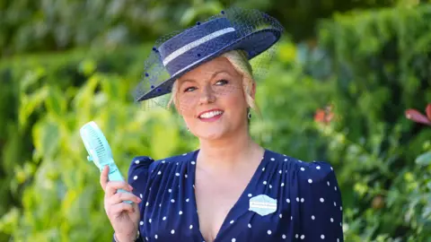 PA Media Natalie Rushdie, who is wearing a blue and white hat and blue dress with white polka dots, is holding a handheld fan in hot weather at Ascot Racecourse.