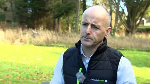 Ryan Williams. He is bald, wearing a black puffer vest with 'Connected Health' written on it and a light blue shirt.