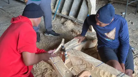 Two men work on carving a tree stump. One man, on the left, who is wearing red overalls and a black beanie, is holding a chisel, the man on the right, dressed in blue, is holding the stump