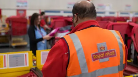 Getty A shot inside a sorting office. A man with short brown hair and glasses has his back to the camera, he is wearing a red Royal Mail issued jacket with a high vis vest over the top saying 'Royal Mail'. In the background are rows of mail trolleys with a woman with dark hair wearing a lanyard looking at parcels.