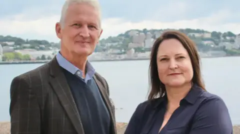 A man in a checked jacket and blue shirt and sweater with white hair and a woman in a blue dress with sea and a town in the background.