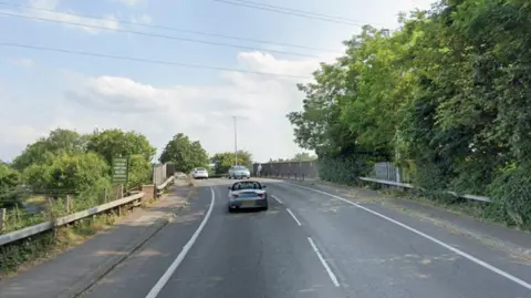 Cars travel over a railway on a road which is bordered by trees either side. There is a footpath over the railway and an opening to some steps on the left hand side.