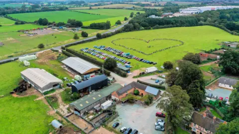 Fordhall An aerial view of a farm, with buildings in the foreground, and people making the shape of a heart in a field to the top of the picture.