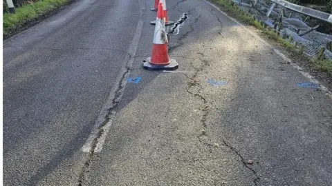 A tarmac road with large cracks with traffic cones along it. Some fencing is falling off the side of the road.