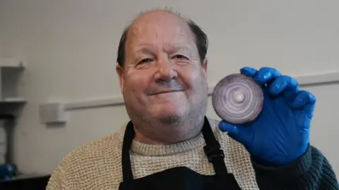 John Fairhall/BBC Vince wears a grey jumper with a black apron on top. He is holding up half an onion and showing it to the camera. His hand has a blue disposable glove on it.
