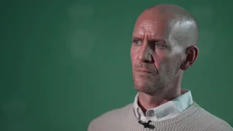 A side profile of Dan Barr, 41, who has blue eyes, a bald head and light stubble, as he looks into the camera wearing a beige jumper over a white shirt.
