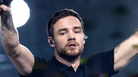 Getty Images Liam Payne, wearing a black top, holds his arms aloft while performing ion stage n 2019. He has short brown hair and a cropped brown beard and moustache