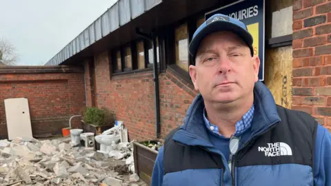 John Devine/BBC Andy Simmonds, 45, wearing a blue baseball cap, blue body warmer over a blue top with a blue and white chequered shirt underneath. He is standing in front of a corner of the old brick pub with a large pile of debris and rubble to the left, including white porcelain toilets and bricks.