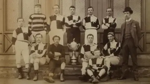 Arthur Wharton Foundation The 12-man football team standing and sitting alongside the Cleveland Challenge Cup. Arthur Wharton is sitting directly to its left with his arms crossed; he wears a darker uniform than his fellow team-mates who are dressed in black-and-white chequered tops and white long-johns. A large silver trophy cup with two large handles is mounted on a plinth.