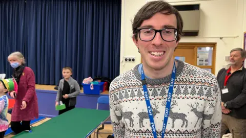 Michael smiles at the camera with his hands held behind his back. He's wearing a white and grey Christmas jumper with reindeer on it. Behind him is a green children's table next to a stage and some children are being guided around. He has hair brown hair which is brushed over and he has squared glasses with black rims.