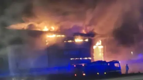 Ian Bennett Fire can be seen coming out of a large building which has the blue and white with yellow Greggs sign on it. Firefighters and fire lorries can be seen and there is a lot of smoke. Its night and there are lights so the picture is not very clear.