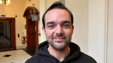 Mickey is smiling at the camera. He is standing in one of the corridors of Belfast City Hall. He has black hair and a beard and is wearing a black jumper with a hood. 