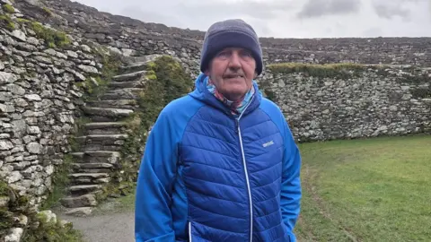 A man in a blue coat and hat stands in the middle of An Grianán of Aileach, a stone ringfort in County Donegal, Ireland.