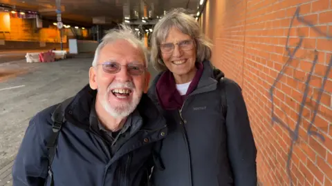 Two older people stand close together beside a long brick wall marked with graffiti. They are wrapped up in multiple layers on a chilly day. They are both smiling. The tunnel roof and lighting reflect softly above them. The ground is a mix of paving and tarmac, giving the area an unfinished feel.