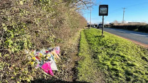 Floral tributes placed along a hedge at the side of Staithes Road where the crash took place. A sign for the village of Preston stands on the grass verge