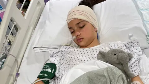 Amelie lying in a hospital bed wearing a white hospital gown. She is unconscious and has her head wrapped in bandages and a pressure cuff on her upper arm. She is also snuggled up with a teddy elephant under her blanket.