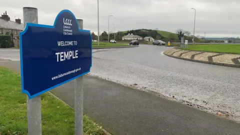 A blue sign that says welcome to Temple, in front of a roundabout.