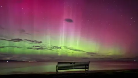 BBC Weather Watchers/Rossi A bench sits in front of the sea in the dark. It is illuminated by a bright green and pink sky, which is dotted with dark clouds. The water reflects from the green lights