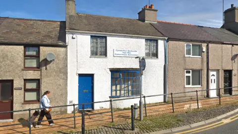 A wide shot the former Ynys Môn Conservative association office in Llangefni, Angelsey. The property has a blue door and glass bay window with a blue window frame. It has the sign that reads in Welsh and English: "Cymdeithas Geidwadol Ynys Môn Conservative Association"