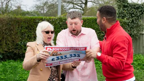 A group of three people standing outdoors. One is wearing a red jumper while the other two are wearing a brown coat and pink shirt and holding a piece of paper which has been styled to look like a cheque.