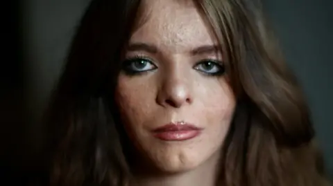 JAMIE NIBLOCK/BBC Amy looks at the camera. She has thick dark brown hair which is parted in the middle and is wavy. Her eyes are green. She is wearing makeup with gloss lipstick on and eye liner with mascara and brown brow liner. The light is caught in the pupils of her eyes and she is in front of a dark background. You can see her face and neck.