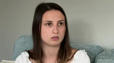 K-Jo sits on a blue sofa in front of a wall. She has shoulder-length, straight, brown hair and wears a necklace and earrings.