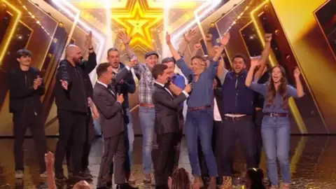 Britain's Got Talent Hawkstone Farmers' Choir on the Britain's Got Talent stage with Ant and Dec. They're all cheering with a gold star behind them.