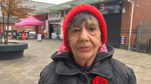 An elderly woman wears a red hat, black coat with a red poppy on and stares into the camera, she is standing in a shopping centre on a cold, autumn day.