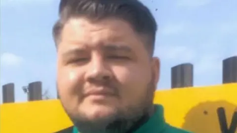 Police Scotland Euan Johnston, a young man with slicked back dark hair and a turquoise green polo shirt, looks into the camera. His eyes are squinting as the sun shines brightly and he has a very slight smile. He is standing against a bright yellow wall.