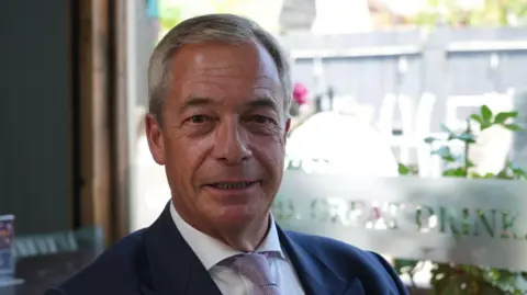 Shaun Whitmore/BBC Nigel Farage smiles for the camera. He has short grey hair and is wearing a blue jacket with a white shirt and purple tie.  