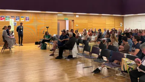 BBC Dozens of people sat inside a school hall for an event. Two people with microphones are sat in front of the audience.