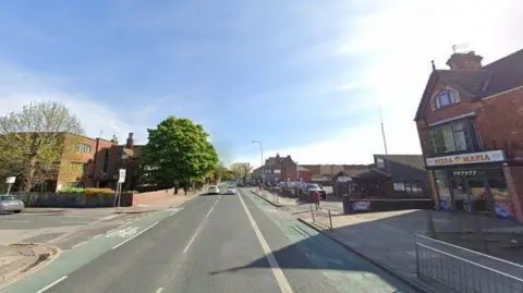 A Google street view of a main road next to several shops including a pizza shop and next to a junction. There are a few cars on the road and green bike lanes either side of the road.