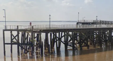 Google Google shot of the wooden pier. It has a safety railing around the walkway, a number of street lamps and a lifebuoy station.