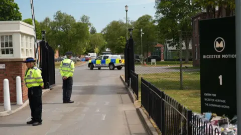 The entrance of Bicester Motion, with open black gates and a lifted barrier. Police officers, one female, one male, guard the entrance, and a police vehicle is in the background. The Bicester Motion sign is on the right, with flower tributes at its base.