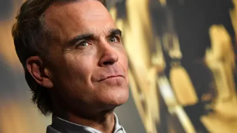 Getty Images Robbie Williams poses for photographers at an event promoting A Better Man, a 2024 film about his career and personal life. A silver shirt collar is visible and he is standing in front of a black and gold backdrop.