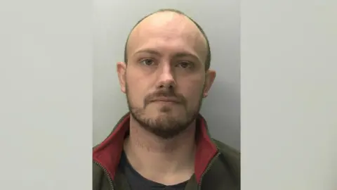 A mugshot of a man with a beard and short brown hair. He is wearing a black top and green and red fleece. The background is grey. 