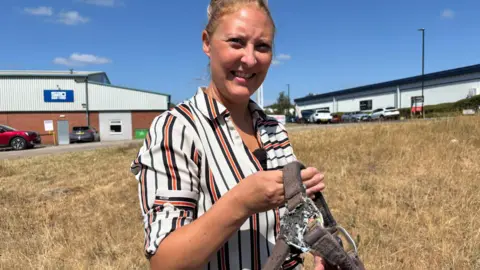 Simon Thake A blonde woman wearing a white striped shirt stands on a grass bank in the sunshine. She's carrying a battered horse collar.