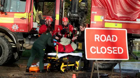 Reuters Rescue workers from the fire and ambulance services lift a person onto a stretcher in front of a red truck on a road which is closed due to large amounts of surface water