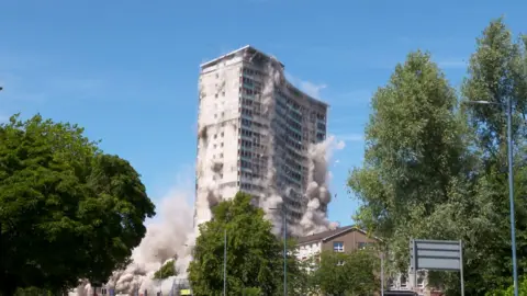 A high-rise tower block mid- demolition. The building is in the middle of collapsing and there are plumes of dust coming out of it. It sits against a blue sky and there are tall trees at either side of the image.