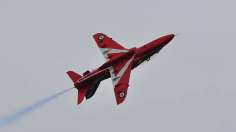 A red jet with white markings flies with blue smoke spraying off its back