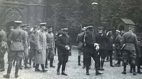 Michelle Bullivant A black and white photo of at least 20 Army officers in a variety of uniforms milling around in front of a college entrance in Cambridge.