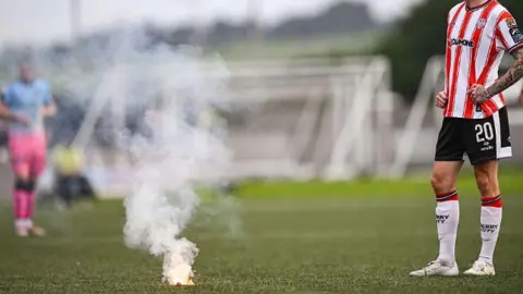Getty Images Shows a flare with smoke coming out of it on a football pitch and a Derry City player on the right in a red and white striped shirt, black shorts, red and white socks and white fooball boots. 