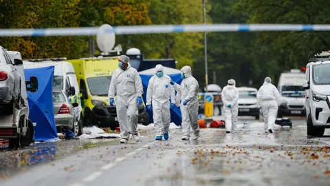 PA Media People walks in hazmat suits - their faces covered with surgical masks and hands covered in blue gloves - walk down a road blocked off by police tape and lined with emergency vehicles