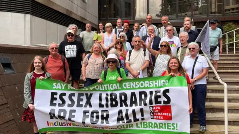 About 30 demonstrators standing on steps outside a large building with a glass front. They are holding a banner saying "Essex libraries are for all".