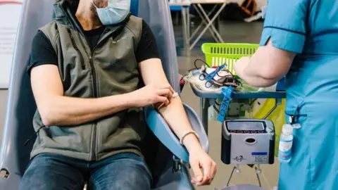 NHS Blood and Transplant A man in a green gilet giving sat in a chair about to donate blood 