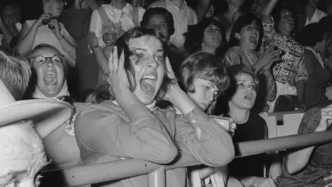 Getty Images Lots of fans stood behind a metal rail screaming because they have seen The Beatles.