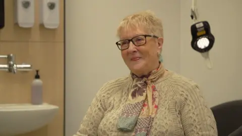 A woman looks slight off camera. She has glasses and short white hair. She is wearing a cream woolly jumper with a colourful scarf. She is sat in what looks like a hospital room.