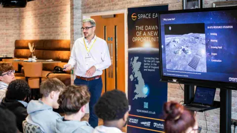 Andrew McCulloch, from Minima, giving a space engineering workshop to students at Suffolk New College. He is wearing a white shirt and navy trousers and addressing a group of students who are sitting down in front of him and a television screen. 